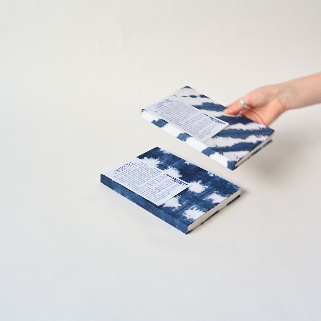 Two hardcover books with blue and white tie-dye patterned covers are shown on a light gray background, with one book being held by a hand.