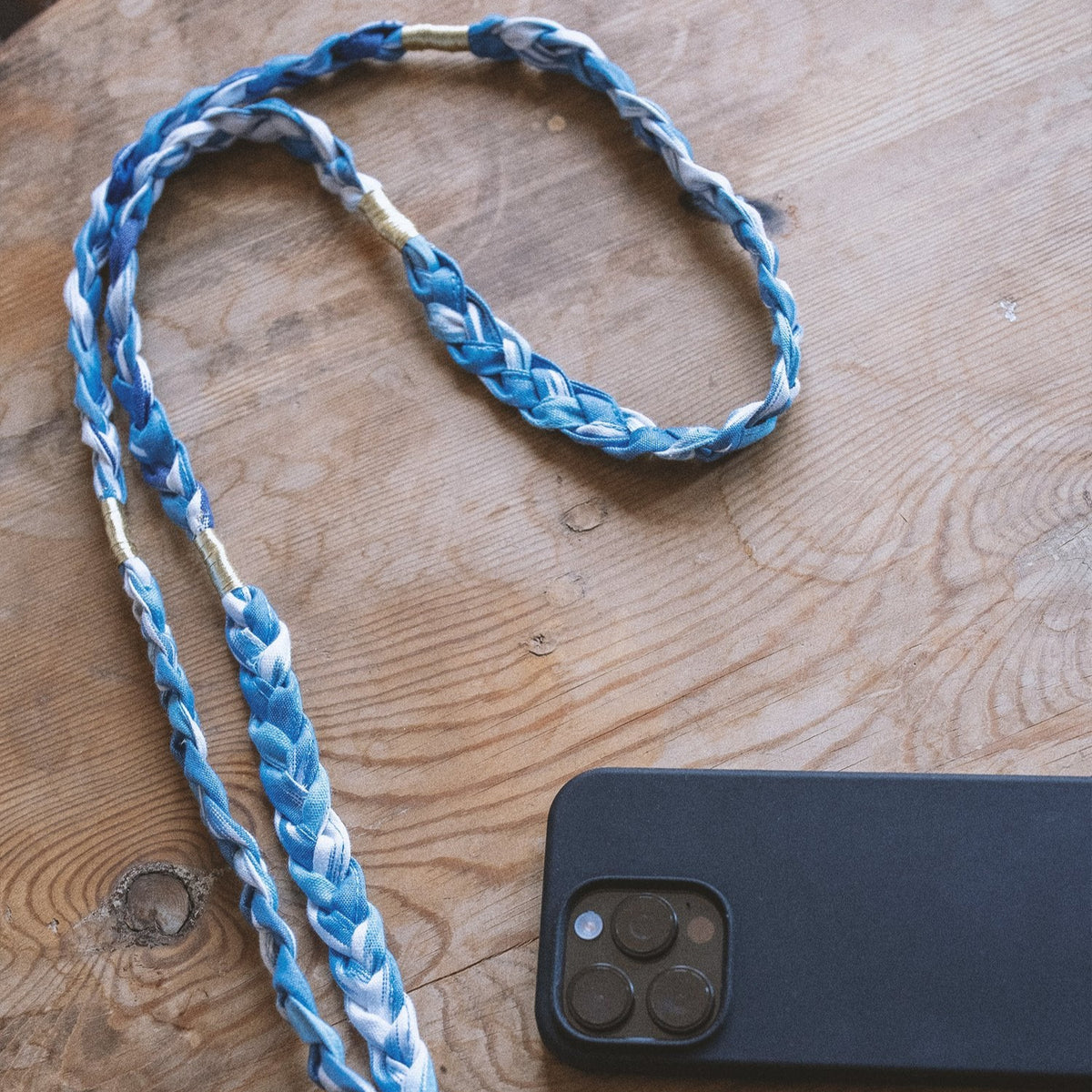Blue and white braided lanyard with a gold tag on a wooden surface next to a black phone.